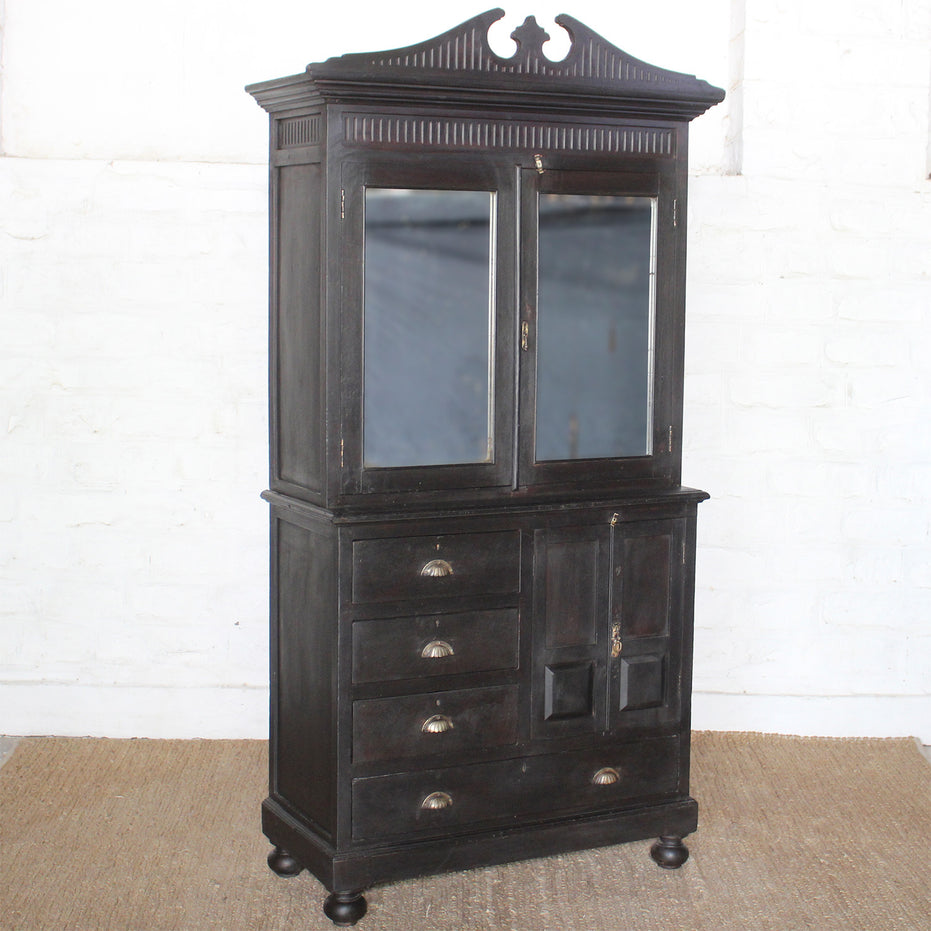 The Rajan Wooden Cabinet by John Robshaw features mirrored double doors on top, four drawers, and two small lower doors for versatile storage; its dark wood finish and round legs add elegance as it stands atop a woven floor mat.