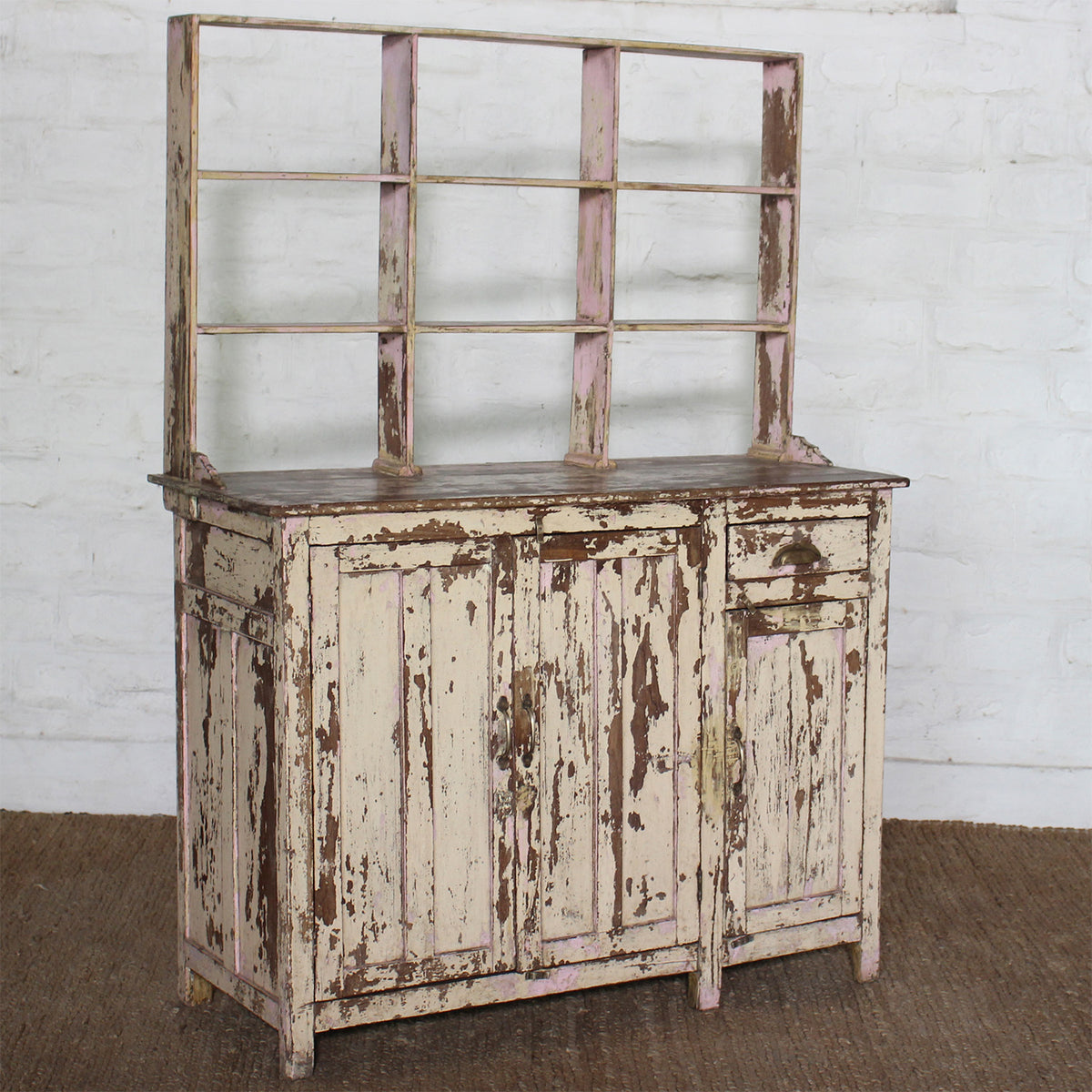 A distressed, authentic furniture piece—the Surya Wooden Side Board by John Robshaw, featuring open shelving above and showcasing chipped white and brown paint—stands against a white brick wall on a brown woven rug.