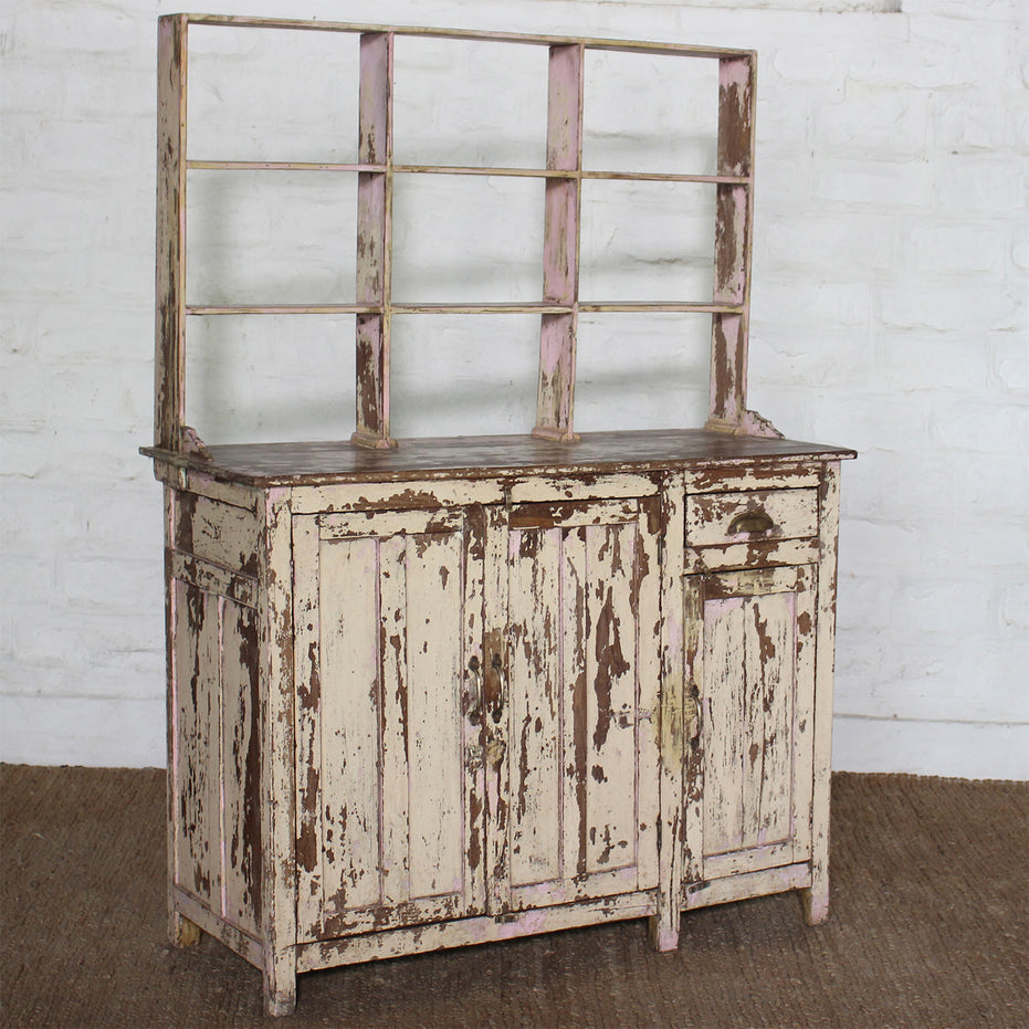 A distressed, authentic furniture piece—the Surya Wooden Side Board by John Robshaw, featuring open shelving above and showcasing chipped white and brown paint—stands against a white brick wall on a brown woven rug.