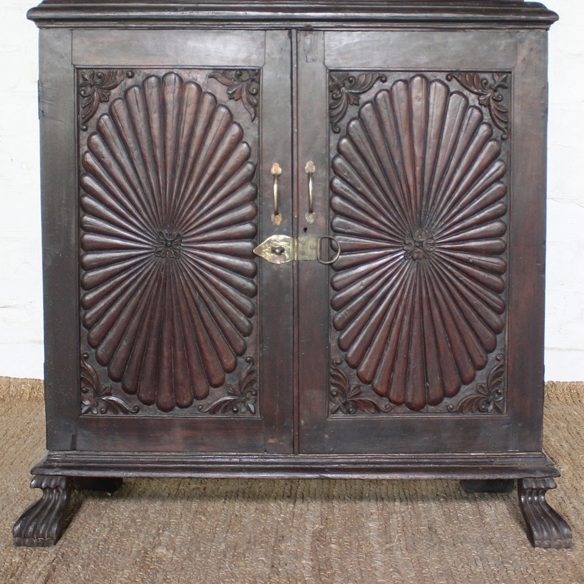 The Mayura Wooden Cabinet by John Robshaw features two ornate, carved sunburst panels with a radial pattern and decorative corners. This dark wooden cabinet sits on clawed feet and is displayed on a woven mat.