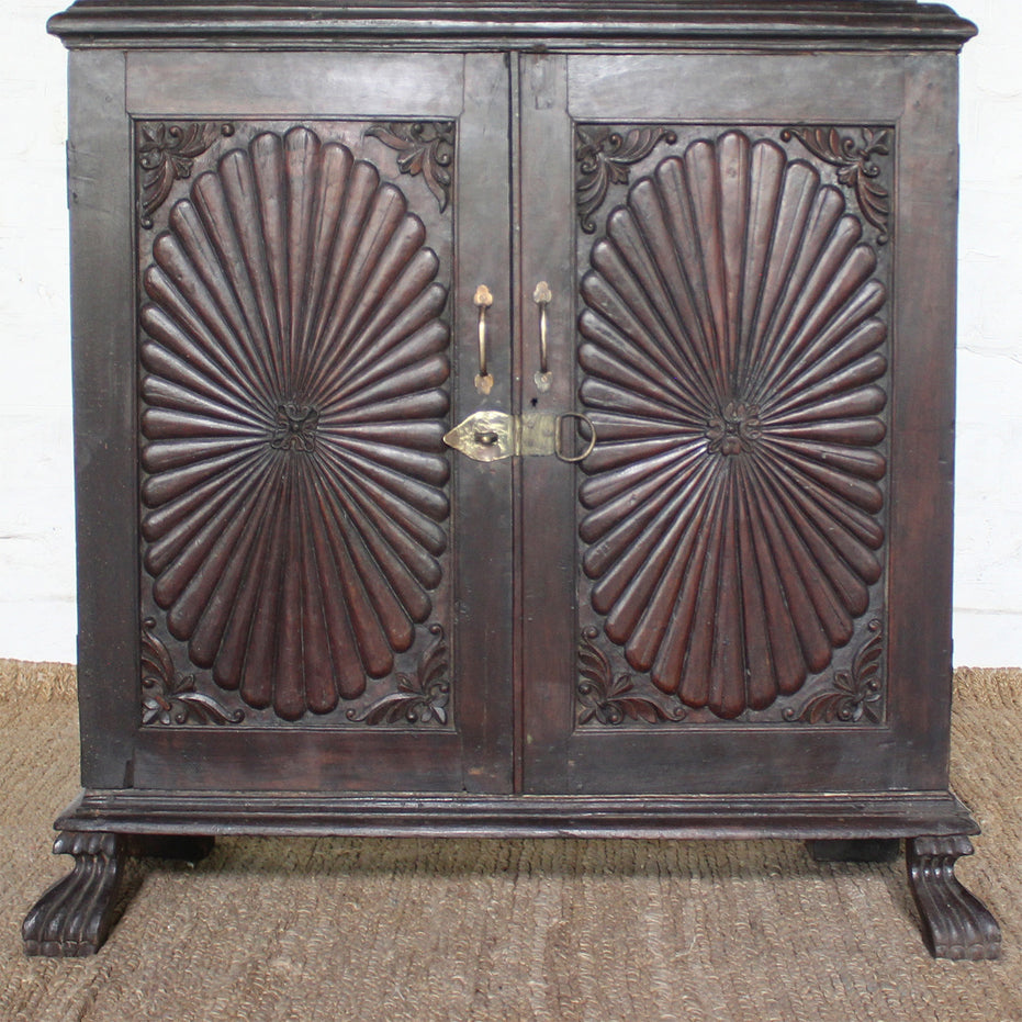The Mayura Wooden Cabinet by John Robshaw features two ornate, carved sunburst panels with a radial pattern and decorative corners. This dark wooden cabinet sits on clawed feet and is displayed on a woven mat.