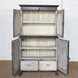 The Mayura Wooden Cabinet by John Robshaw is a vintage dark wood piece featuring four open doors, three shelves, and two drawers with elegant carved sunburst panels. It stands on beautifully carved legs and is showcased against a white brick wall.