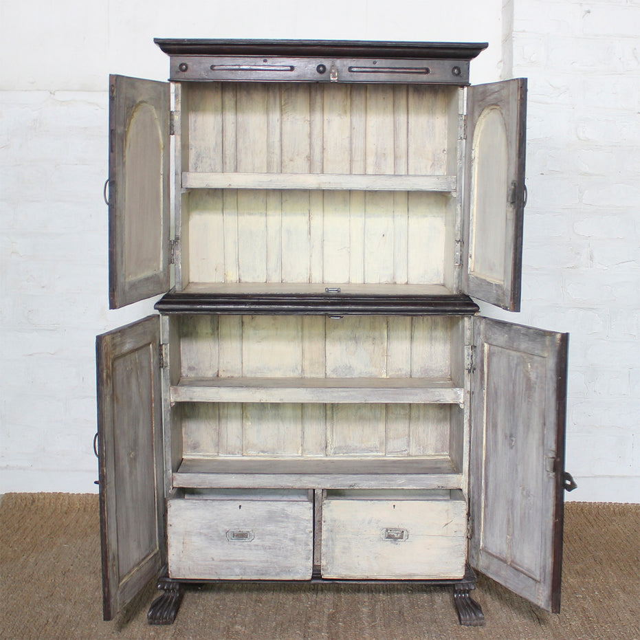 The Mayura Wooden Cabinet by John Robshaw is a vintage dark wood piece featuring four open doors, three shelves, and two drawers with elegant carved sunburst panels. It stands on beautifully carved legs and is showcased against a white brick wall.