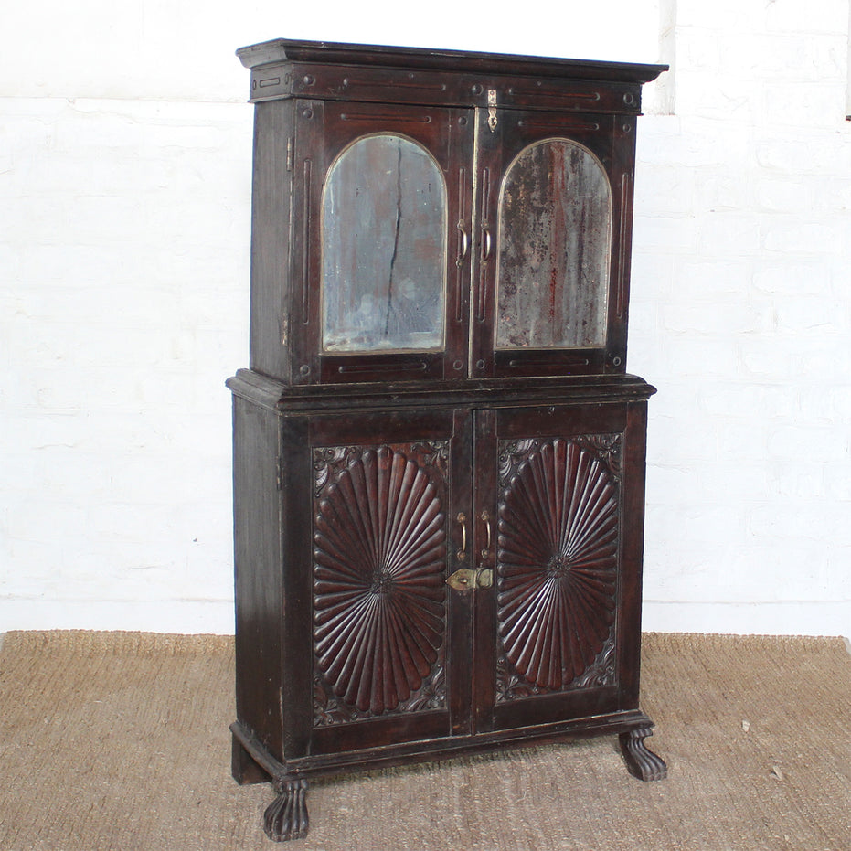 The Mayura Wooden Cabinet by John Robshaw features antique dark wood, mirrored upper doors, intricately carved floral designs on the lower doors, and elegant claw feet, positioned against a white brick wall atop a woven mat.