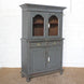The Revati Wooden Cabinet by John Robshaw showcases a dusky blue finish with two arched glass doors on top, two central drawers, and two solid lower doors adorned with elegant brass accents—all supported by round feet and set against a white wall background.