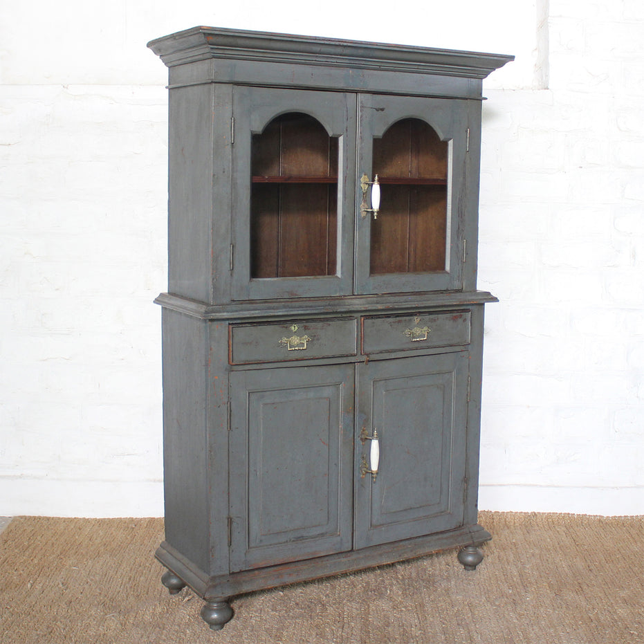 The Revati Wooden Cabinet by John Robshaw showcases a dusky blue finish with two arched glass doors on top, two central drawers, and two solid lower doors adorned with elegant brass accents—all supported by round feet and set against a white wall background.