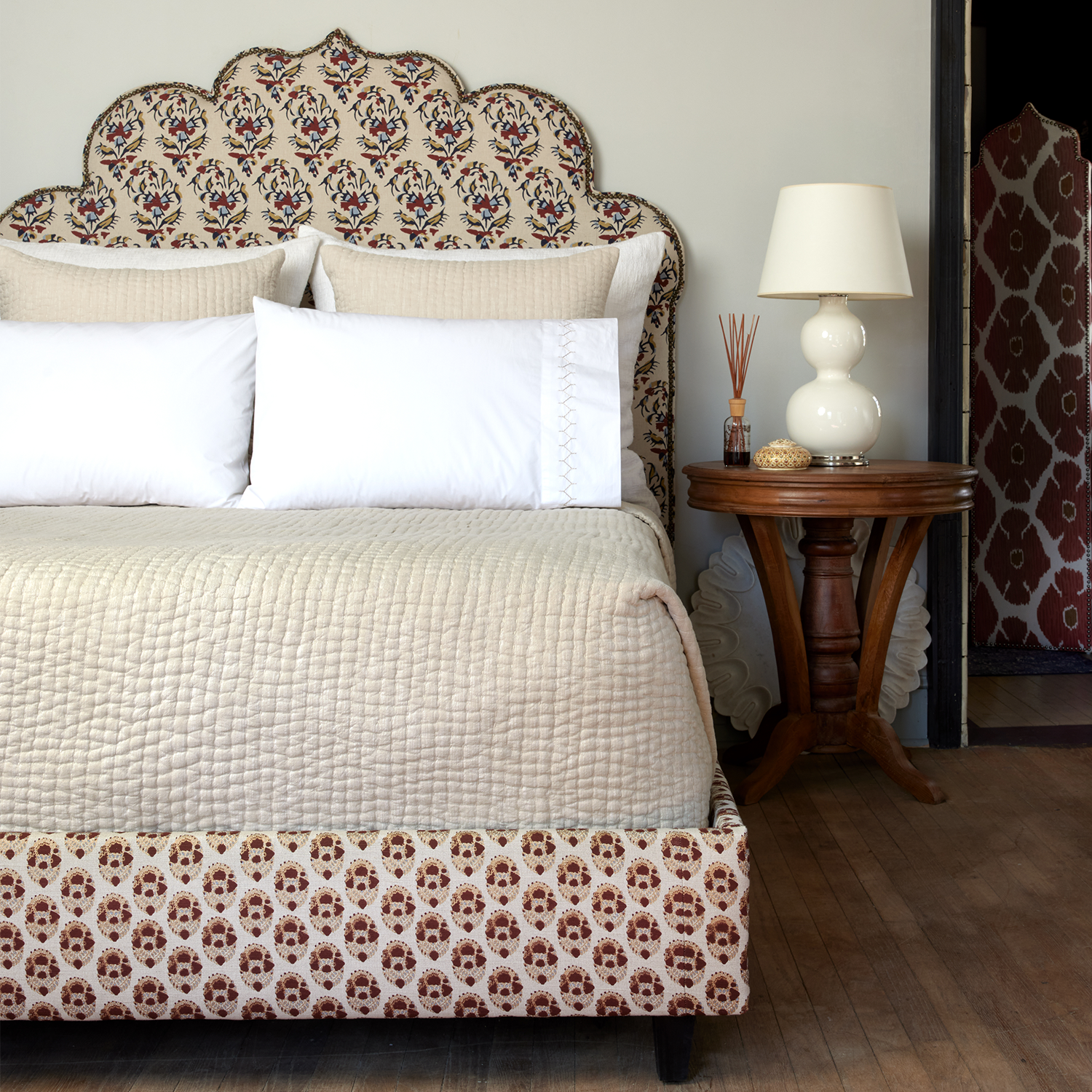 A bed with a patterned headboard covered in cotton chambray coverlets and adorned with hand stitching, featuring the Vivada Sand Woven Quilt by John Robshaw.